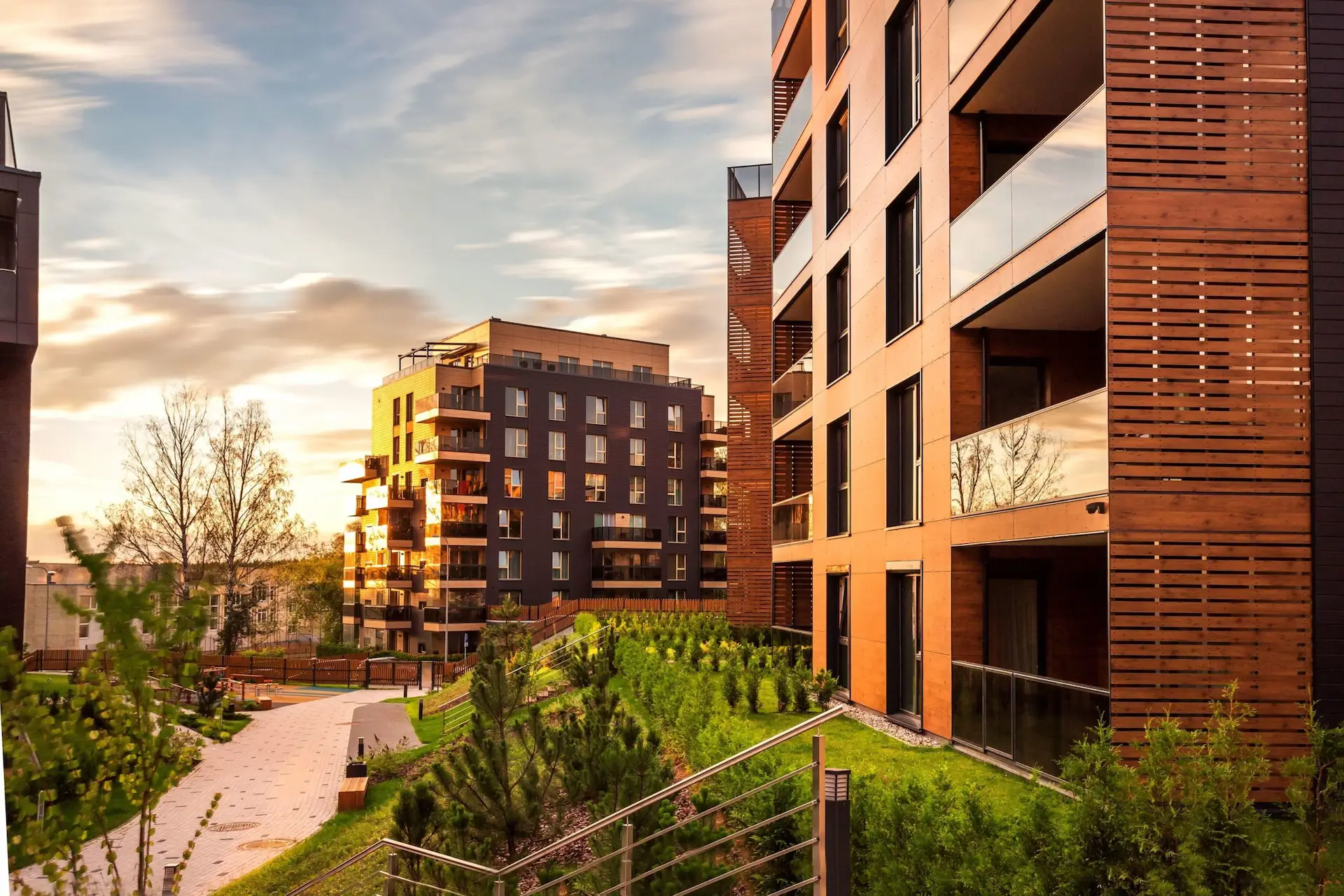 Modern residential apartment complex at sunset. The buildings feature a contemporary architectural design with brown and dark grey facades, wooden pergola elements, and glass balconies. A landscaped pathway winds between the buildings, lined with young evergreen trees and well-maintained grass areas. The warm golden light of sunset reflects off the buildings' surfaces, creating a welcoming atmosphere. The development showcases sustainable urban design with its integration of green spaces and pedestrian-friendly walkways.